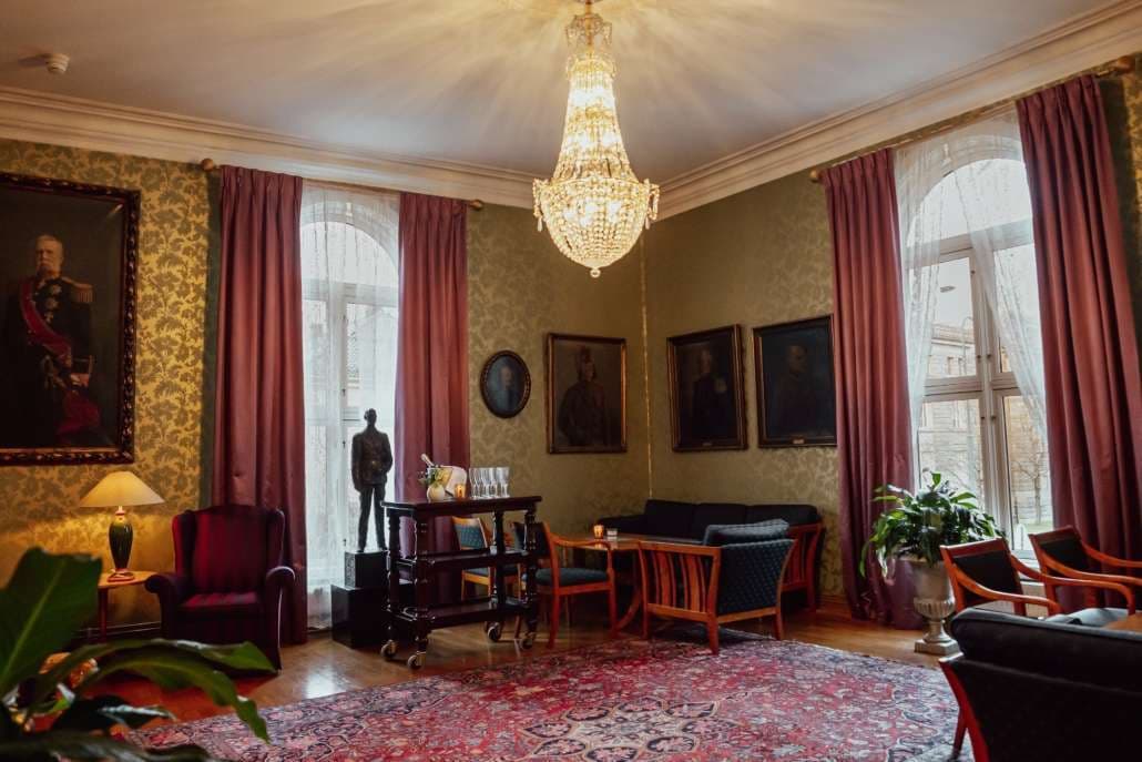 Salong med gullfarget tapet, røde gardiner og lysekrone.